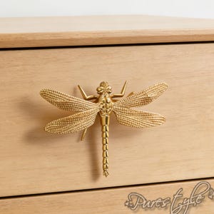 May include: A gold-colored dragonfly drawer pull mounted on a light-colored wooden cabinet. The dragonfly has detailed wings and a segmented body. The cabinet has two drawers and a simple design.