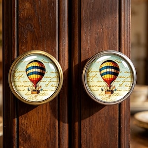 May include: Two round cabinet knobs featuring a vintage-style hot air balloon illustration. The design displays a colourful balloon with blue, yellow, and red stripes against a background of cursive script text, encased in a metallic circular frame.