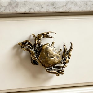 Puede incluir: Un tirador de mueble de metal color latón con forma de cangrejo, con patas y pinzas detalladas. El cangrejo está montado en un frente de cajón de color crema. El texto "Purestyle" es visible en la esquina inferior derecha en cursiva.