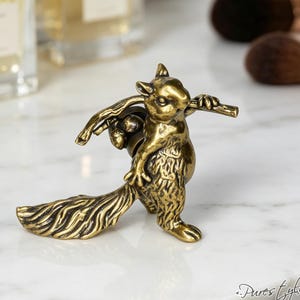 May include: A brass-toned metal cabinet knob shaped like a standing squirrel. The squirrel is depicted with a textured fur finish, holding a small branch across its shoulders with two acorns hanging from it. The piece has a metallic antique gold finish.
