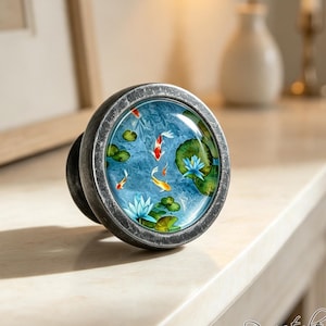May include: A round cabinet knob with a dark, weathered metallic finish and a glass insert. The insert features a vibrant illustration of orange and white koi fish swimming among green lily pads and blue lotus flowers on a textured blue background.