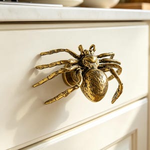 May include: A brass-colored metal cabinet knob shaped like a spider with eight detailed legs and a textured, rounded body. The spider is mounted onto a cream-colored kitchen drawer, featuring a metallic finish that reflects light across its surface.