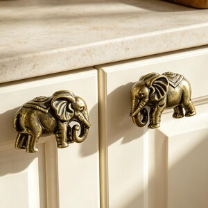Puede incluir: Dos pomos de armario de latón envejecido con forma de elefante, cada uno con texturas detalladas y una trompa curvada, montados en dos puertas de armario de cocina de color blanco roto con un acabado liso y diseño de panel rectangular.