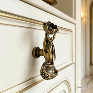 May include: A brass-toned metal drawer knob shaped like a hand holding a lotus flower. The hand is positioned vertically, with the fingers grasping the stem of the flower at the top. The base of the knob features an intricate, textured pattern.