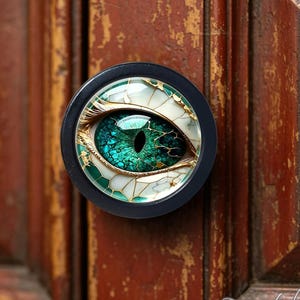 Pode incluir: Um puxador de móvel circular com um gráfico detalhado de um olho de réptil com uma íris verde vibrante e uma pupila preta. O olho é rodeado por um padrão rachado em branco e dourado, envolto num acabamento liso e brilhante com uma moldura preta.