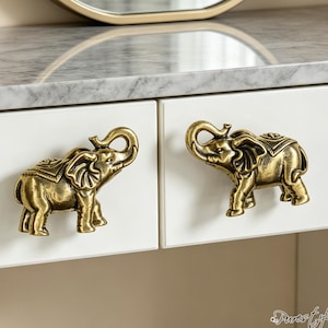 Può includere: Due pomelli per armadietti in metallo color ottone antico a forma di elefante con le proboscidi sollevate, montati su frontali di cassetti bianchi. Le figure degli elefanti presentano texture dettagliate e sono rivolte l'una verso l'altra.