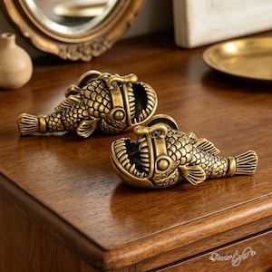 以下が含まれることがあります： アンコウの形をした真鍮色の金属製キャビネットノブ2個。各ノブには、質感のある鱗模様、目立つ背びれ、歯が見える開いた口が特徴です。仕上げはアンティークゴールドカラーで、暗い色合いの陰影が施されています。