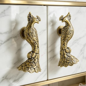 Puede incluir: Dos tiradores de armario de latón antiguo con forma de aves estilizadas con plumaje y crestas detalladas, montados en puertas de armario de mármol blanco con vetas grises, enmarcados por un borde de metal dorado fino en los bordes.