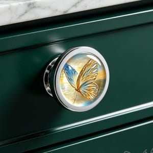 May include: A round cabinet knob with a polished silver metal frame and a circular insert featuring a detailed illustration of a butterfly with blue and gold wings against a soft, glowing background, mounted on a dark green drawer front.