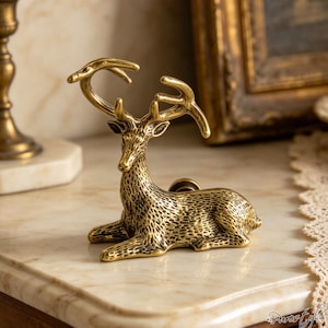 May include: A brass-coloured metal cabinet knob shaped like a reclining deer with detailed fur texture and prominent antlers. The deer is depicted in a resting position with its head turned slightly to the side, featuring a circular mounting base on its back.