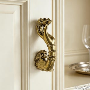 May include: A brass-coloured cabinet handle shaped like an arm extending from a base, with the hand holding a lotus flower. The arm features detailed wrist bands and a textured, curved mounting plate. The handle is attached to a cream-coloured cabinet door.