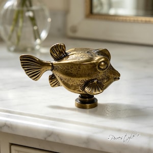 May include: A brass-toned metal cabinet knob shaped like a pufferfish. The knob features a textured, weathered surface with detailed fins, a prominent eye, and a small mouth. It is mounted on a circular base, showcasing a metallic finish.