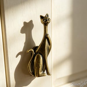 Peut inclure: Un bouton de placard en métal couleur laiton en forme de chat stylisé en position assise avec un cou et une queue allongés. La figurine de chat présente une finition métallique lisse avec des détails antiques sombres, fixée sur une surface en bois clair.