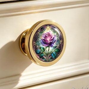 May include: A round gold-toned cabinet knob featuring a central circular inlay with a detailed illustration of a purple rose. The rose is surrounded by intricate green leaves and decorative dark purple and teal patterns within a stained glass style frame.