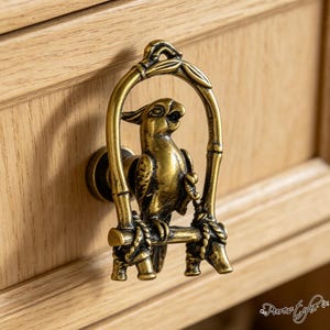 May include: A brass-toned metal drawer knob featuring a three-dimensional parrot perched on a swing. The parrot is positioned inside an arched frame that resembles bamboo stalks, with rope-like details securing the perch to the frame structure.