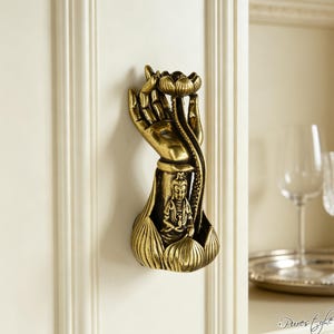 May include: A metallic gold-toned wall-mounted decorative object featuring a sculpted hand holding a lotus flower above a small seated figure. The base is shaped like a lotus blossom, and the entire piece has an antique finish with dark accents.