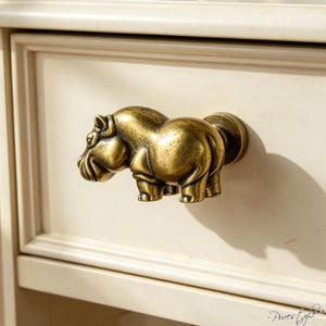 May include: A brass-toned metal drawer knob shaped like a hippopotamus. The figure is depicted in profile, facing left, with a smooth, rounded body and a circular mounting base attached to a cream-colored wooden drawer front with a frame detail.