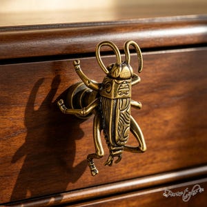 May include: A brass-toned metal drawer knob shaped like a grasshopper. The insect figure features detailed etched lines on its body, wings, and legs, with two prominent antennae extending upward. The knob is mounted on a circular base.