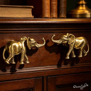 May include: Two metallic gold-toned elephant-shaped drawer pulls mounted on a dark wooden cabinet. Each elephant figure is depicted in profile with a raised trunk, textured skin details, and a polished finish, serving as functional hardware.
