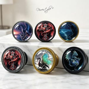 May include: Six round cabinet knobs featuring glass domes with dragon illustrations. The designs include red dragons on crosses, blue dragons over water, and teal-winged dragons. The knobs have metallic frames in silver, gold, and black finishes.