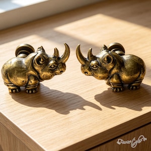 May include: Two brass-colored metal cabinet knobs shaped like rhinoceroses. Each knob features a detailed rhinoceros figure with a prominent horn, textured skin, and a circular mounting base on the back for attachment to furniture or cabinetry.