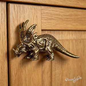 May include: A brass-coloured metal cabinet knob shaped like a triceratops dinosaur. The figure features a textured, detailed surface with a prominent frill, three horns, and a long tail, mounted horizontally on a light-coloured wooden cabinet door.