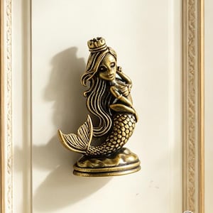 May include: A brass-toned metal cabinet knob shaped like a mermaid wearing a crown. The mermaid has long flowing hair, a detailed scaly tail, and is positioned on a rounded base. The hardware is mounted on a white panel with gold trim.