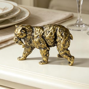 Puede incluir: Una figura de oso en tono dorado metálico con acabado texturizado, representada en posición de caminar con un pequeño pez en la boca. La escultura presenta detalles de pelaje marcados y una forma sólida y compacta como objeto decorativo.