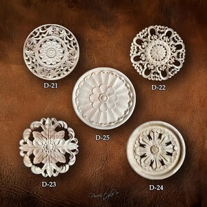 Peut inclure: Cinq éléments décoratifs circulaires en bois sculpté avec des motifs floraux et ornementaux. Chaque pièce est d'un style différent, étiquetée D-21, D-22, D-23, D-24 et D-25. Le fond est une surface marron texturée.