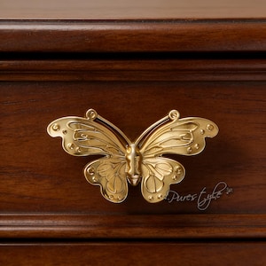 May include: A gold-coloured butterfly-shaped drawer pull fixed to a dark brown wooden surface. The butterfly design features detailed wing patterns and a textured finish, enhancing the furniture's aesthetic.