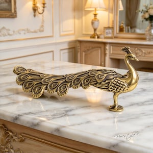 May include: A metallic gold-toned peacock figurine featuring an elongated, fanned tail with detailed feather patterns. The bird stands on a single pedestal base, showcasing a polished finish with darkened accents in the recessed areas of the design.