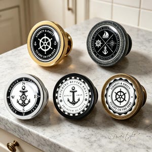 Può includere: Cinque pomelli circolari per mobili con disegni nautici. I pomelli presentano grafiche in bianco e nero di timoni, ancore e bussole con testo che include Liberty & Amity, Smooth Sailing e Building Better Boats. Le basi sono oro, argento, nero e bronzo.