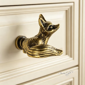 May include: A brass-toned metal cabinet knob shaped like a fox with a textured finish, featuring a detailed head and a curled tail. The knob is mounted on a cream-colored drawer front. The text Pures tyle is visible in the bottom right corner.