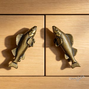 May include: Two antique brass-toned metal cabinet pulls shaped like fish, featuring detailed scale textures, fins, and eyes. The fish are oriented diagonally, mounted on light-colored wooden cabinet doors with visible grain patterns.