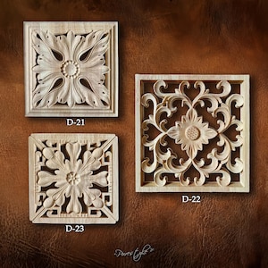 May include: Three square, wooden decorative panels with carved floral designs. The panels are light-colored and feature intricate details, including leaves and a central flower. Each panel is labeled with a "D-" number.