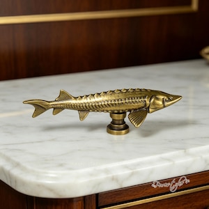 Peut inclure: Un bouton de meuble en métal couleur laiton en forme d'esturgeon. Le poisson présente des écailles, des nageoires et un museau pointu détaillés, monté sur une base cylindrique. La finition a une patine sombre de style ancien avec des reflets.