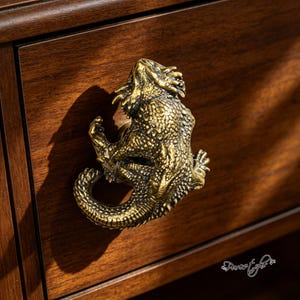 Op de afbeelding: Een messingkleurige meubelknop in de vorm van een baardagame met gedetailleerde schubben, stekels en een gekrulde staart. De knop is bevestigd op een donkerbruine houten ladefront en heeft een driedimensionaal reptielontwerp.