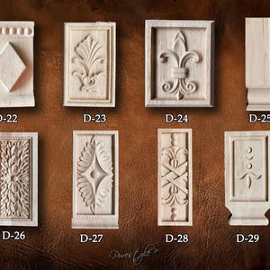 May include: A collection of eight unfinished wooden decorative architectural elements. Each piece features intricate carved designs, including floral patterns, geometric shapes, and a fleur-de-lis. The pieces are labeled D-22 through D-29.