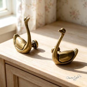 Puede incluir: Dos ganchos de pared de metal en tono latón antiguo con forma de cisne. Cada gancho presenta un cuello curvado y un cuerpo redondeado, con una base de montaje circular en la parte posterior. El acabado tiene una textura metálica desgastada.
