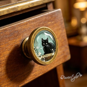 May include: A round cabinet knob with an antique brass finish featuring a glass-covered insert. The insert displays a black cat sitting among white flowers against a light blue wooden background, mounted on a wooden drawer front.