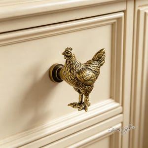 Puede incluir: Un tirador de mueble de metal en tono latón con forma de gallina de pie. La figura detallada presenta plumas texturizadas y una cresta definida, montada sobre una base circular fijada a un frente de cajón color crema con marco.
