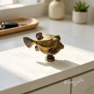 May include: A brass-toned metal cabinet knob shaped like a pufferfish. The knob features a textured, weathered finish with detailed fins, a round eye, and a small mouth. It is mounted on a circular base, designed for attachment to furniture or doors.