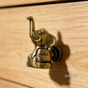 Puede incluir: Un pomo de mueble de metal en tono latón con forma de elefante sentado con la trompa levantada. La figura del elefante presenta un acabado metálico liso con zonas hundidas oscurecidas para resaltar los detalles esculpidos de las orejas y el cuerpo.