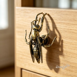 May include: A brass-toned metal cabinet knob shaped like a wasp with detailed wings, legs, and segmented body. The insect-shaped hardware is mounted onto a light-colored wooden drawer front, casting a distinct shadow to the right of the object.