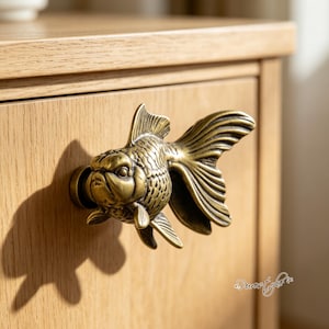 May include: A brass-toned metal drawer knob shaped like a goldfish with detailed scales and fins. The knob is attached to a light-colored wooden drawer front. The fish figure has a prominent tail and fins, with a metallic finish that reflects light.