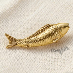 May include: A gold-colored fish-shaped brooch. The brooch has detailed scales and fins, with a textured surface. The fish is lying on a cream-colored, textured surface.