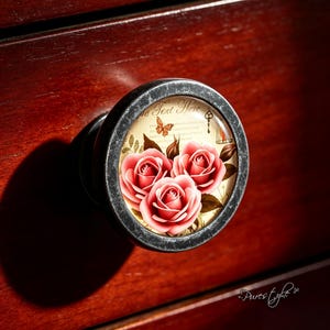 May include: A round cabinet knob with a distressed dark metal frame featuring a central glass insert. The insert displays three pink roses, a small orange butterfly, a key, and the text "The Text Here" on a cream-colored background.