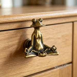 May include: A brass-toned metal drawer knob shaped like a frog sitting in a meditative pose with its legs crossed and hands resting on its knees. The frog has a smooth, metallic finish with darkened recessed areas to highlight its physical features.