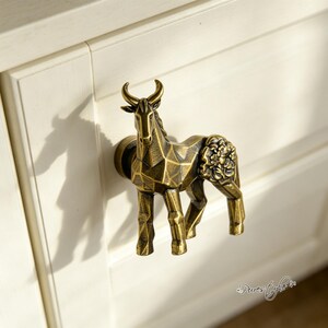 May include: A brass-toned metal cabinet knob shaped like a standing animal with horns, featuring a geometric faceted body and a detailed floral relief pattern on the hindquarters, mounted on a circular base attached to a white cabinet door.