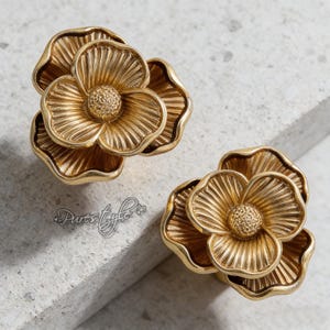 Könnte beinhalten: Zwei goldfarbene Schrankknöpfe in Blumenform. Jeder Knopf hat detaillierte Blütenblattdesigns und eine strukturierte Mitte. Die Knöpfe sind aus Metall und werden wahrscheinlich zur Möbeldekoration verwendet. Die Knöpfe befinden sich auf einer hellgrauen Oberfläche.